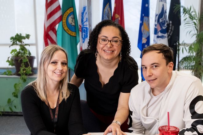 Three people at table with flags in background. 